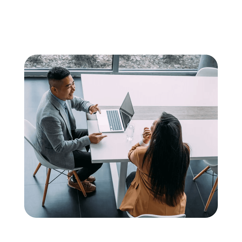 Two professionals having a discussion at a table with a laptop in a modern office setting with large windows.